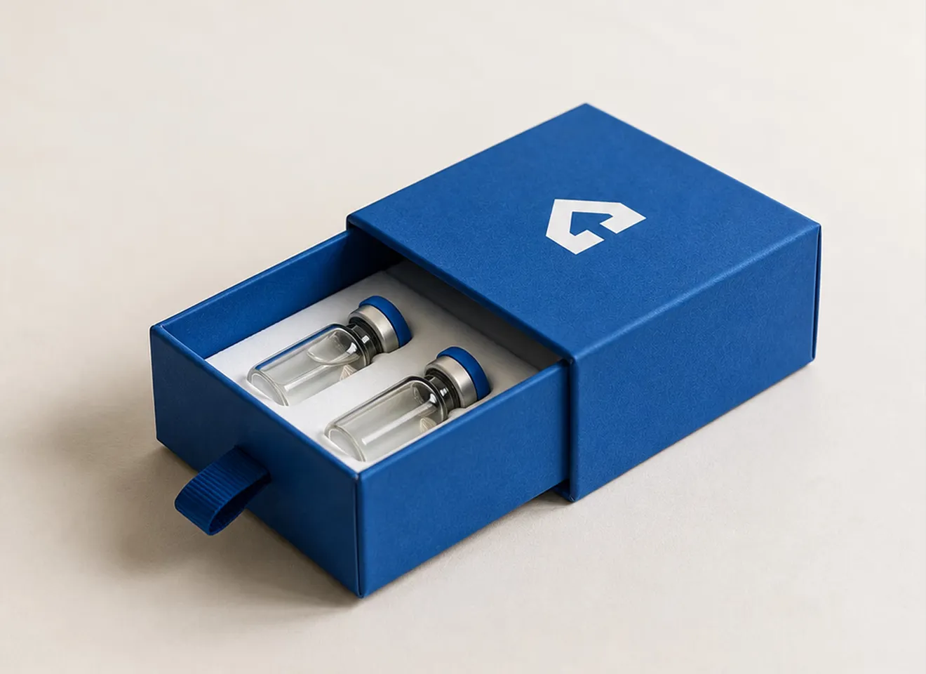 A blue slide-out drawer box with a grosgrain pull tab, opened to show two glass vials seated upright in a white foam insert.