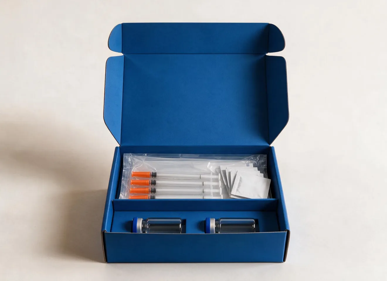 An open blue mailer box holding a protocol kit — four orange-capped syringes sealed in a clear sleeve and a stack of white sachets in the upper tray, with two capped glass vials seated in cut-outs below.